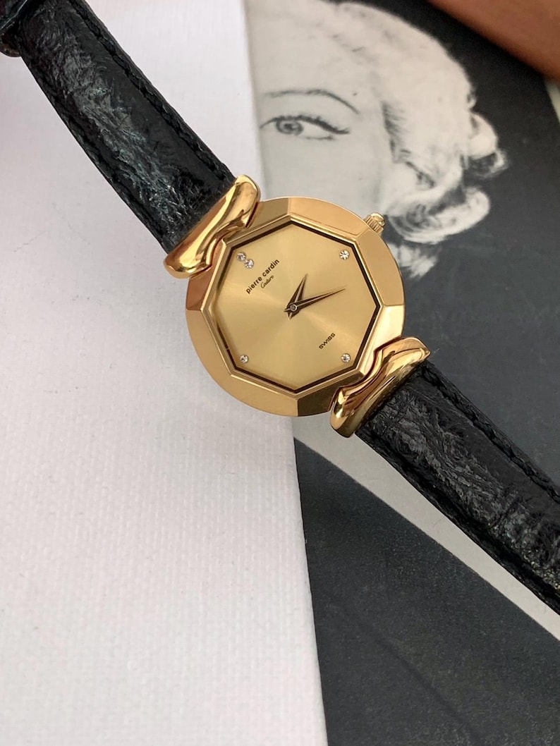 May include: A gold-tone Pierre Cardin watch with a black leather band. The watch face is octagonal and features a gold-tone dial with diamond accents.