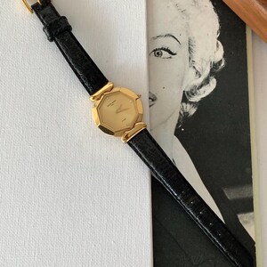 May include: A gold-tone wristwatch with a black leather band. The watch face is octagonal and has a gold-tone bezel with small diamonds. The watch is on a white surface.