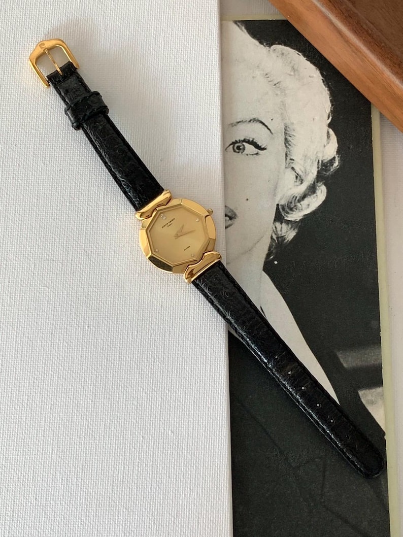 May include: A gold-tone wristwatch with a black leather band. The watch face is octagonal and has a gold-tone bezel with small diamonds. The watch is on a white surface.