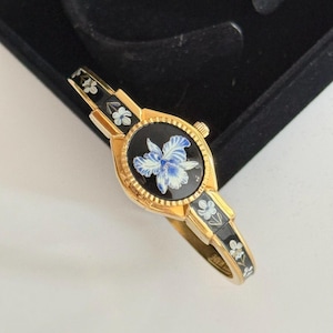 Puede incluir: Un reloj de pulsera rígida en tono dorado con una esfera ovalada sobre fondo negro y un diseño floral azul y blanco. La banda está decorada con paneles de esmalte negro con pequeñas flores blancas y hojas verdes, a juego con el motivo floral.