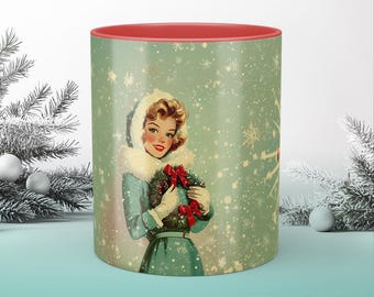May include: A green ceramic mug with a red rim features a vintage illustration of a woman in a blue and white winter outfit holding a wreath with red berries. The background is a light green with a snowflake pattern.