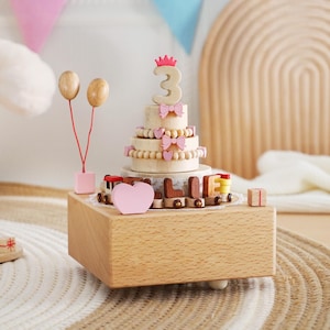 Peut inclure: Bo&icirc;te &agrave; musique en bois en forme de g&acirc;teau d'anniversaire avec le chiffre 3, des n&oelig;uds roses et un train avec le nom "ELLIE". Comprend des ballons en bois et un c&oelig;ur rose. Le g&acirc;teau est pos&eacute; sur une bo&icirc;te en bois.