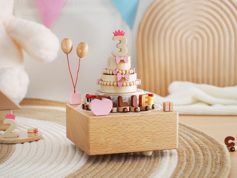 Peut inclure: Bo&icirc;te &agrave; musique en bois en forme de g&acirc;teau d'anniversaire avec le chiffre 3, des n&oelig;uds roses et un train avec le nom "ELLIE". Comprend des ballons en bois et un c&oelig;ur rose. Le g&acirc;teau est pos&eacute; sur une bo&icirc;te en bois.