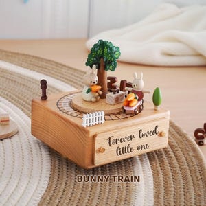 Peut inclure: Bo&icirc;te &agrave; musique en bois sur le th&egrave;me du train de lapins. Elle pr&eacute;sente un lapin avec une carotte, un arbre et un train transportant des carottes. La bo&icirc;te est grav&eacute;e de "Forever loved little one" et "BUNNY TRAIN".