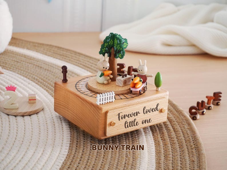 Peut inclure: Bo&icirc;te &agrave; musique en bois sur le th&egrave;me du train de lapins. Elle pr&eacute;sente un lapin avec une carotte, un arbre et un train transportant des carottes. La bo&icirc;te est grav&eacute;e de "Forever loved little one" et "BUNNY TRAIN".