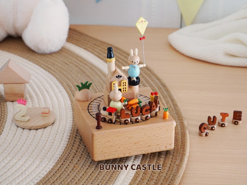 Peut inclure: Bo&icirc;te &agrave; musique en bois sur le th&egrave;me du ch&acirc;teau de lapin. La bo&icirc;te pr&eacute;sente un train avec le nom "LUCA" et un lapin tenant un cerf-volant. Le texte "BUNNY CASTLE" est imprim&eacute; sur le devant.