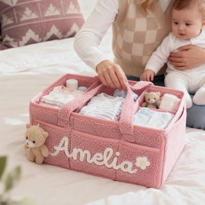 Op de afbeelding: Een roze baby-organizer mand met meerdere compartimenten, gevuld met babykleding, een teddybeer en benodigdheden voor de baby. De naam "Amelia" is op de voorkant geborduurd, samen met een bloem. De mand heeft handvatten.