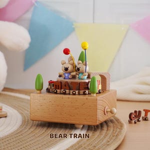 Peut inclure: Bo&icirc;te &agrave; musique en bois en forme de train avec deux oursons tenant des ballons. Le train porte le mot "MATE". La bo&icirc;te &agrave; musique est marron clair avec des arbres verts. Le mot "CUTE" est &eacute;pel&eacute; en petites lettres en bois.