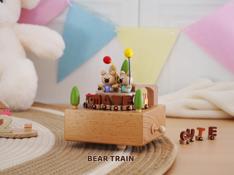 Peut inclure: Bo&icirc;te &agrave; musique en bois en forme de train avec deux oursons tenant des ballons. Le train porte le mot "MATE". La bo&icirc;te &agrave; musique est marron clair avec des arbres verts. Le mot "CUTE" est &eacute;pel&eacute; en petites lettres en bois.