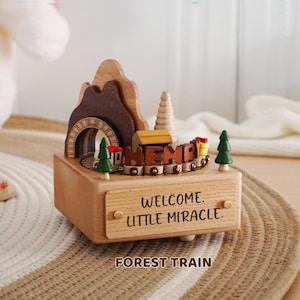 Peut inclure: Bo&icirc;te &agrave; musique en bois avec un train, un tunnel et un design de montagne. La bo&icirc;te porte les inscriptions "WELCOME LITTLE MIRACLE" et "FOREST TRAIN". Comprend deux petits arbres verts et un train avec le mot "HEMP".