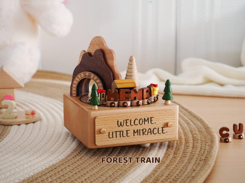 Peut inclure: Bo&icirc;te &agrave; musique en bois avec un train, un tunnel et un design de montagne. La bo&icirc;te porte les inscriptions "WELCOME LITTLE MIRACLE" et "FOREST TRAIN". Comprend deux petits arbres verts et un train avec le mot "HEMP".