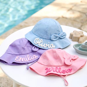 Pode incluir: Três chapéus de sol pastel, azul claro, lavanda e rosa, cada um com um laço e o nome bordado: "Olivia", "Emilia" e "Willow". Os chapéus estão numa mesa branca perto de uma piscina.