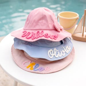 Könnte beinhalten: Stapel von drei pastellfarbenen Sonnenhüten. Der obere Hut ist rosa mit dem Namen "Willow" in Rosa gestickt. Der mittlere Hut ist hellblau mit "Olivia" in Weiß gestickt. Der untere Hut ist rosa mit "Chloe" in Regenbogenfarben gestickt.