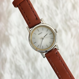 May include: A Seiko wristwatch featuring a round silver-tone case with a white dial, thin black hour markers, and slender hands. The watch is attached to a smooth, stitched brown leather strap with a matching brown leather keeper loop.