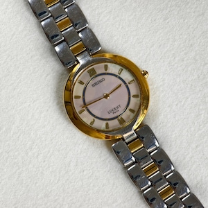 Könnte beinhalten: Eine Seiko Lucent Paris Armbanduhr mit goldfarbener Lünette und einem zweifarbigen silber-goldenen Armband. Das Zifferblatt der Uhr ist hellrosa mit goldenen Zeigern und römischen Ziffern. Die Uhr wird auf einer weißen Oberfläche präsentiert.