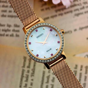 May include: A rose gold-tone Seiko solar wristwatch featuring a round case with a crystal-encrusted bezel. The watch has a mother-of-pearl dial with pink stone hour markers, gold-tone hands, and a fine rose gold-tone mesh metal link strap.