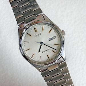 May include: A silver-toned Seiko Chronos wristwatch featuring a round white dial with gold-colored hour markers and hands. The watch includes a day-date window at the three o'clock position and a matching silver-toned metal link bracelet strap.