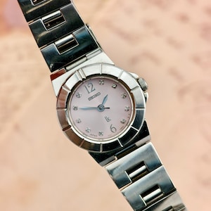 May include: A silver-tone stainless steel Seiko Lukia wristwatch featuring a pale pink dial with diamond hour markers, the numbers 12 and 6, and silver hands. The watch has a round bezel with notched detailing and a matching link bracelet.