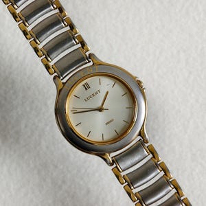Op de afbeelding: Een Lucent polshorloge met een zilver- en goudkleurige metalen band en kast. De wijzerplaat van het horloge is wit met gouden wijzers en markeringen. Het woord "LUCENT" is op de wijzerplaat gedrukt. De horlogeband is ongeveer 18 cm lang.