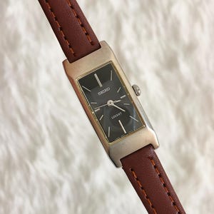 May include: A rectangular Seiko Lucent wristwatch featuring a black dial with silver-tone hour markers and hands. The watch has a silver-tone metal case and a brown leather strap with visible stitching along the edges and a side crown.