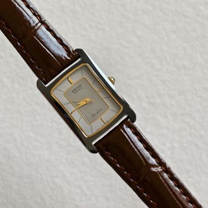 Puede incluir: Un reloj Seiko Quartz Exciline rectangular con esfera plateada y detalles dorados. El reloj tiene una correa de cuero marrón. La esfera muestra el logotipo de Seiko y la palabra "Exciline".