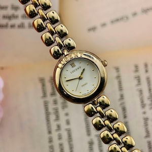 May include: A gold-tone wristwatch with a round white dial featuring the text Lucent and Seiko. The watch has a polished gold-tone metal link bracelet with rounded square segments. The bezel is engraved with the word Lucent at the top position.