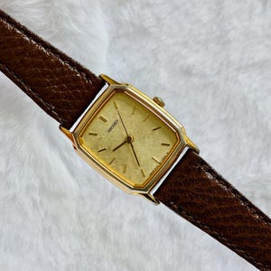 Puede incluir: Un reloj de pulsera rectangular Seiko con caja dorada y esfera dorada texturizada con finas manecillas e índices negros. El reloj está sujeto a una correa de cuero marrón oscuro con una textura granulada visible y bordes cosidos.