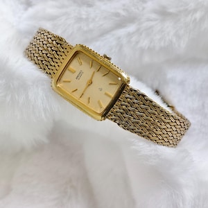 Puede incluir: Un reloj Seiko Quartz dorado con una esfera rectangular y una correa de metal trenzado. La esfera del reloj es de color oro claro con marcadores de hora y agujas negras. La correa es de color oro a juego, sobre un fondo blanco y esponjoso.