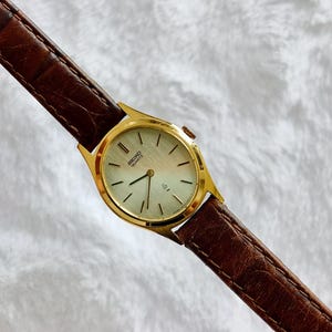Può includere: Un orologio da polso Seiko Quartz color oro con quadrante rotondo di colore chiaro, indici dorati sottili e lancette affusolate. L'orologio è fissato a un cinturino in pelle marrone scuro con una finitura testurizzata effetto coccodrillo.