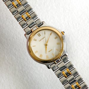 May include: A two-tone wristwatch featuring a silver and gold-tone metal link bracelet. The round watch face has a white dial with gold-tone Roman numeral hour markers, gold-tone hands, and the text Lucent and Seiko printed on the dial.