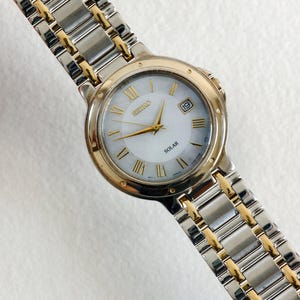 Puede incluir: Un reloj de pulsera solar Seiko con caja redonda en tonos plata y oro y esfera blanca texturizada. El reloj cuenta con marcadores de hora en números romanos dorados, manecillas doradas y una ventana de fecha en la posición de las tres.