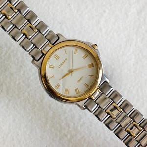 Op de afbeelding: Een zilver- en goudkleurig Lucent horloge met een crèmekleurige wijzerplaat en gouden wijzers. Het horloge heeft Romeinse cijfers en een zilver- en goudkleurige schakelband. Een klassiek horloge.