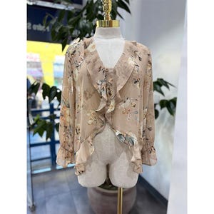 May include: A beige floral blouse with a V-neck and ruffled trim. The long-sleeved top features a delicate floral pattern in shades of beige, pink, and green. The blouse is displayed on a mannequin.