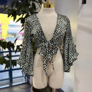 May include: A cropped, long-sleeve top with a tie front. The top features a black, brown, and green animal print pattern on a light blue background. The sleeves have a flared, ruffled design. The top is displayed on a mannequin.