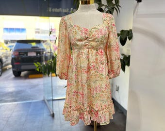May include: A floral print dress with long sleeves and a square neckline. The dress features a ruffled hem and a fitted bodice. The fabric has a cream background with pink and green floral patterns. The dress is displayed on a mannequin.