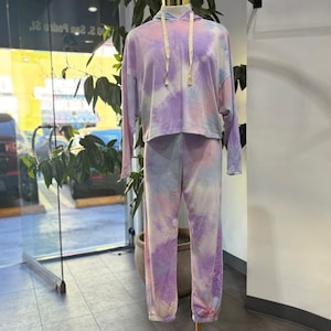 Può includere: Un completo da relax tie-dye nelle tonalità del viola, rosa e blu. Il set comprende una felpa con cappuccio con coulisse e pantaloni della tuta abbinati. L'outfit è esposto su un manichino in un ambiente di negozio.
