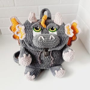 May include: A handmade gray crochet dragon backpack with orange and yellow wings, light pink accents, and green eyes. The backpack has a top handle, adjustable straps, and a metal clasp closure. The dragon's face is detailed with horns and a snout.