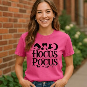 May include: A pink t-shirt with the words "Hocus Pocus" in black, along with witch-themed graphics. The shirt is being worn by a person in blue jeans. The background is a brick wall and greenery.
