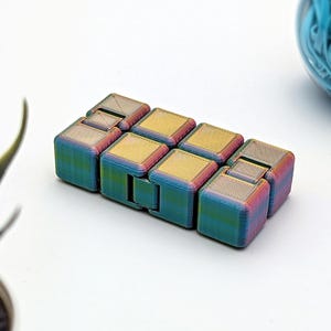 Könnte beinhalten: Ein mehrfarbiges Fidget-Spielzeug aus kleinen Würfeln, die gedreht und geklappt werden können. Das Spielzeug besteht aus einem metallischen Material mit einem regenbogenfarbenen, irisierenden Finish.