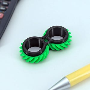 Könnte beinhalten: Ein schwarzes und grünes Fidget-Spielzeug mit Zahnrad-Design. Das Spielzeug ist aus Kunststoff und hat eine glatte, abgerundete Oberfläche.