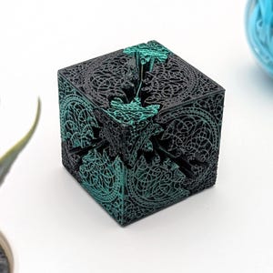 Könnte beinhalten: Ein schwarzer und türkisfarbener 3D-gedruckter Würfel-Puzzle mit komplizierten geometrischen Mustern. Der Würfel ist aus Kunststoff und hat eine glatte Oberfläche.