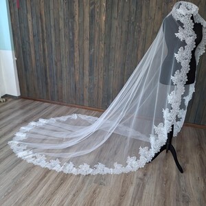 May include: A white tulle wedding veil with a delicate lace trim. The veil is displayed on a mannequin and is draped over a wooden background.
