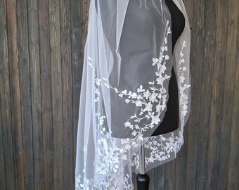 May include: A white bridal veil with delicate floral embroidery along the edges. The veil is made of sheer fabric and is draped over a black mannequin, showcasing the intricate design and texture. The background is a wooden wall.