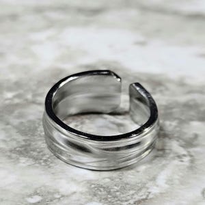 May include: A silver ring with a textured, wavy design. The ring is open on one side, allowing it to be adjusted to fit different finger sizes.