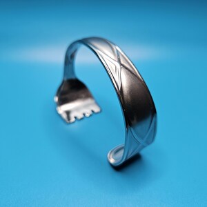 May include: A silver metal bracelet with a geometric pattern. The bracelet is curved and has a textured surface.