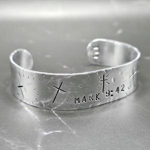May include: Silver bracelet with a hammered texture and the words "Mark 9:42" and a cross engraved on it.