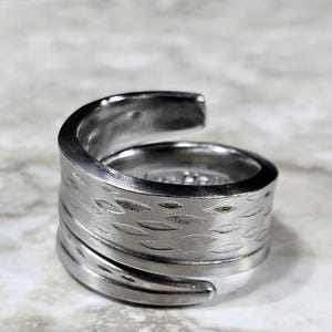 May include: A silver-colored ring with a wrap-around design. The ring features a textured band with a repeating leaf pattern. The ring is made of a shiny, metallic material.