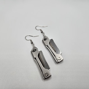 May include: A pair of silver-colored earrings in the shape of miniature utility knives. Each earring features a small blade and a handle with a screw detail. The earrings hang from silver-colored fish hook ear wires.