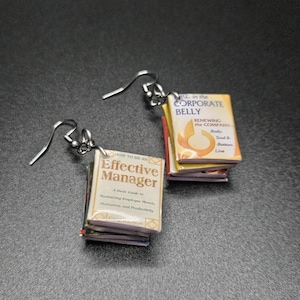 May include: Silver-tone dangle earrings featuring miniature book charms. One earring displays "HOW TO BE AN Effective Manager" and the other reads "KE in the CORPORATE BELLY". The earrings are a unique accessory for book lovers.