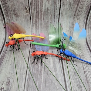 May include: A collection of colorful dragonfly garden stakes. The dragonflies are in various colors, including red, yellow, green, and blue, with translucent wings. Each dragonfly is attached to a green wire stem.